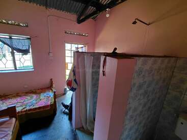 This is a small pink room with a single bed, tiled shower cubicle, curtained window, mosquito net, and metal roof, lit by natural daylight.