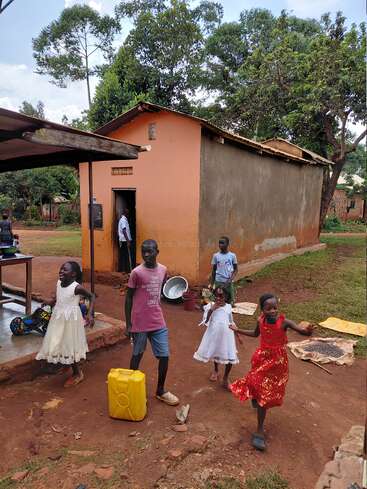 Several children in colorful clothes play and walk near a small house with mud walls. Trees and greenery surround the area, creating a lively, rural atmosphere.
