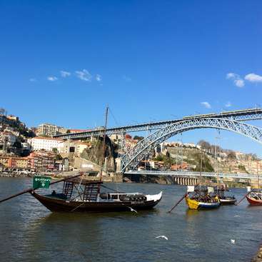 The image depicts a serene river scene with boats and a bridge in the background, set against a backdrop of buildings on a hillside under a clear blue sky.
