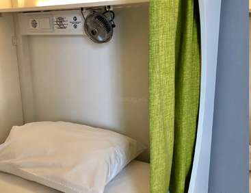 The image depicts a compact, white room with a bed, pillow, and green curtain, featuring a small fan on the wall and a white electrical outlet.