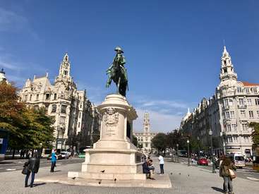 The image depicts a city square with a statue of a man on a pedestal, surrounded by people, buildings, and vehicles, set against a blue sky.
