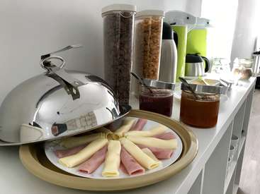 The image depicts a breakfast buffet with a silver serving dish, ham and cheese rolls, and various condiments on a white counter against a white wall.