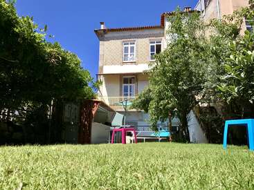 The image depicts a serene backyard scene with a two-story house, lush trees, and vibrant furniture, set against a clear blue sky.