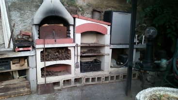 This image shows an outdoor kitchen area with a wood-fired oven, storage shelves for firewood, various tools, a table, and surrounding rustic decor.