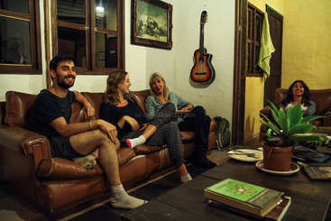 L'image représente un groupe de personnes réunies dans une pièce, entourées d'une table et de chaises, avec une grande fenêtre à l'arrière-plan, probablement un salon ou une salle à manger.
