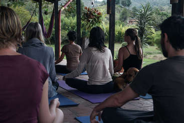L'image représente un groupe de personnes pratiquant le yoga ou la méditation, assises sur le sol, les jambes croisées, dans un cadre extérieur serein. L'atmosphère est paisible et apaisante.