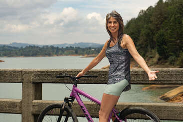 A woman stands beside a purple bicycle on a bridge, smiling. Behind her is a scenic lake, lush trees, distant hills, and a partly cloudy sky.