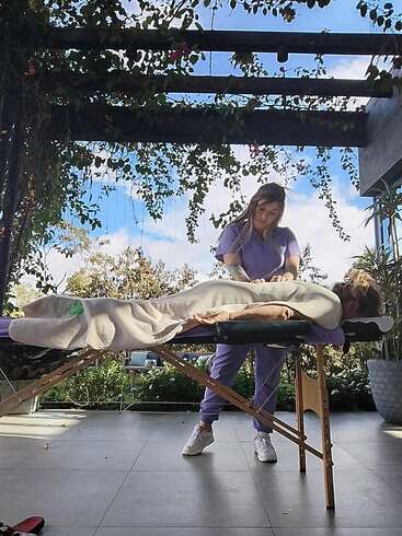 A woman receives a relaxing outdoor massage on a portable table under a pergola with hanging plants, while another woman in purple scrubs provides the treatment.