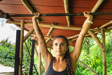 A woman with braided hair is performing a pull-up exercise outdoors under a bamboo shelter, surrounded by lush greenery and natural sunlight, appearing focused and determined.