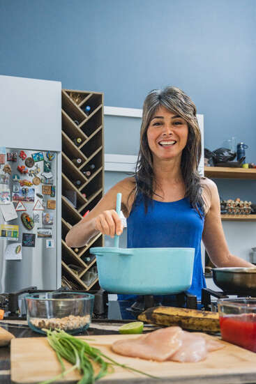 A smiling woman in a blue top cooks in a modern kitchen, stirring a pot. Ingredients like chicken, green onions, and sauce are on the counter.