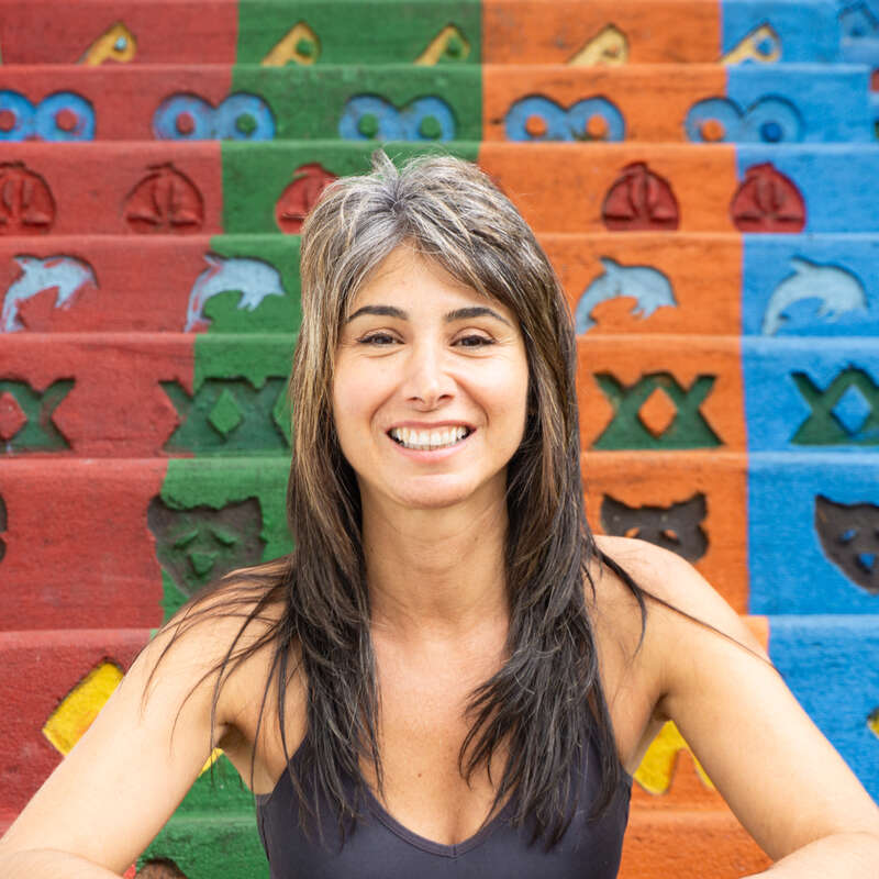A smiling woman with dark hair and light highlights poses in front of vibrant, colorful stairs decorated with playful animal and geometric shapes in red, green, orange, and blue.