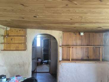 This image shows a rustic room with wooden ceiling and shelves, plastered walls, an arched doorway, and visible clutter. Natural light enters from an adjoining room.