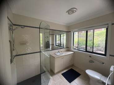 This bathroom features a glass shower, large mirror, single vanity sink, toilet with grab bar, tiled floor, and a big window overlooking a green garden.
