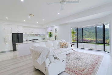This bright, modern living room features a white sectional sofa, cozy blanket, elegant rug, open kitchen, large windows, and stunning views of lush greenery.