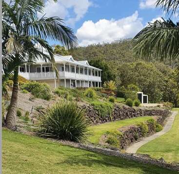 A beautiful white house with a large wraparound porch sits on a hillside, surrounded by lush greenery, palm trees, manicured gardens, and a stone retaining wall.