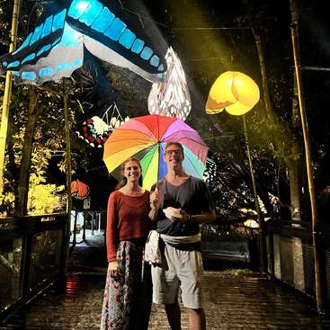 The image depicts a couple standing on a bridge at night, surrounded by large butterfly sculptures and holding a rainbow umbrella.