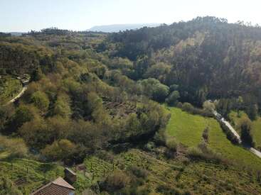 This aerial image shows a lush, green valley with dense forests, scattered fields, winding roads, and gentle hills under clear, bright daylight, creating a serene landscape.