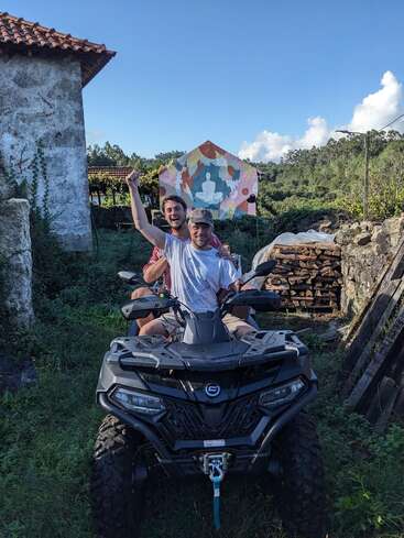 The image depicts two men riding an ATV in a rural setting, with one man raising his arm in celebration or excitement.