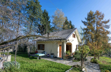 Ein kleines gelbes Haus mit Ziegeldach steht in einem üppigen, grünen Garten, umgeben von Bäumen, Büschen und einem klaren, blauen Himmel über uns. Friedliche Umgebung.