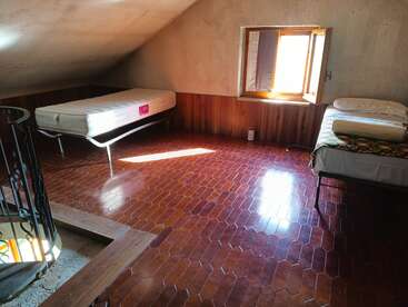 Dieses Bild zeigt ein einfaches Schlafzimmer im Dachgeschoss mit zwei Einzelbetten, einer Wendeltreppe, sechseckigen braunen Fliesen, Holzvertäfelung und Sonnenlicht, das durch ein Fenster fällt.