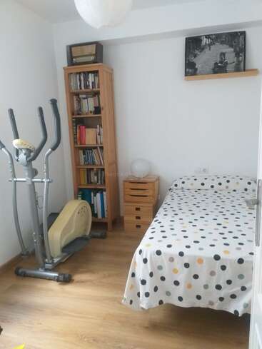 Cette chambre confortable comprend un lit simple avec une couverture à pois, une étagère, un vélo d'appartement, des tiroirs en bois, une lampe, une radio vintage et une photo en noir et blanc.