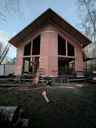 Ein halbfertiges Haus mit Holzrahmen und Holzverkleidung, umgeben von Baumaterialien, Leitern und Geräten. Ein Hund sitzt im schlammigen Vordergrund zwischen Bäumen.
