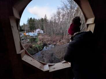 Jemand in Winterklamotten guckt aus einem runden Holzfenster raus auf eine Waldlandschaft, mit einem Haus, einem Wohnmobil und einem Bach im Hintergrund.