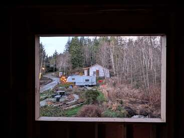 Das Bild zeigt einen Wohnwagen, ein kleines Haus, einen Picknicktisch und eine waldige, rustikale Landschaft mit winterlichen Bäumen und gewundenen Pfaden, eingerahmt von einem Fenster.