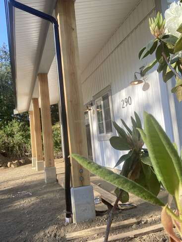 Cette photo montre un bâtiment blanc avec des colonnes en bois sur le porche, des lampes d'extérieur, le numéro « 290 », des plantes au premier plan et un sol en terre battue, le tout pris en plein soleil.