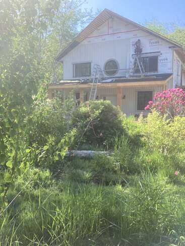 Une maison en construction recouverte d'une membrane Tyvek, avec des échelles appuyées contre ses murs, entourée de plantes et d'arbres à la végétation luxuriante. Un soleil éclatant et un ciel dégagé au-dessus.