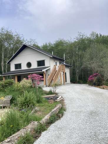 Ein modernes zweistöckiges Haus mit einer Kiesauffahrt, umgeben von üppigem Grün und blühenden Blumen, mit einer Holztreppe und einem runden Fenster im Obergeschoss.