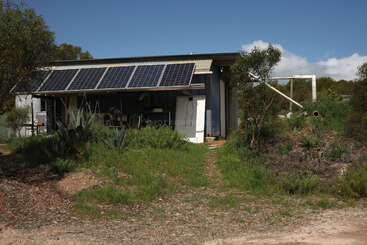 Un petit bâtiment rustique au toit incliné est équipé de panneaux solaires. Entouré de verdure et de buissons, il se dresse sous un ciel bleu ensoleillé et s'inscrit dans une démarche de développement durable.