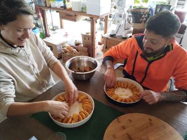 Deux personnes sont assises à une table de cuisine, disposant des tranches de pêches dans des plats de cuisson ronds, souriant et appréciant le processus de cuisson ensemble dans une cuisine confortable et encombrée.