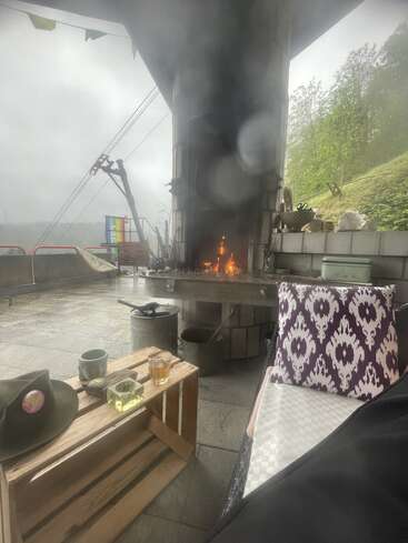 Un cadre extérieur chaleureux avec une cheminée allumée, une table en caisses sur laquelle sont posés des verres, un chapeau, un coussin à motifs, un temps brumeux et une végétation luxuriante en arrière-plan. Une atmosphère paisible.