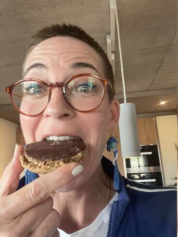 Une personne aux cheveux courts et portant des lunettes sourit alors qu'elle s'apprête à manger un biscuit enrobé de chocolat dans une cuisine moderne ; elle porte des boucles d'oreilles bleues et une veste.