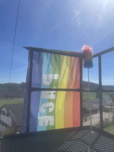 Un drapeau arc-en-ciel aux couleurs vives portant l'inscription « PACE » est accroché à une balustrade métallique à l'extérieur. Un plumeau arc-en-ciel est accroché à proximité, sous un ciel bleu éclatant et ensoleillé, avec un paysage rural en arrière-plan.