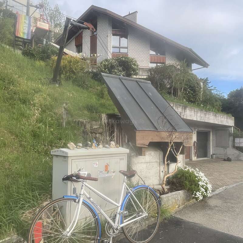 Un vélo blanc est garé devant un coffret électrique orné de pierres peintes. Derrière, on aperçoit une maison située sur une colline verdoyante, entourée de végétation.