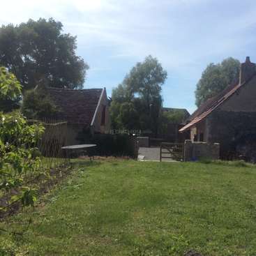 L'image représente une scène rurale sereine avec une cour herbeuse, une petite maison et un grand arbre, sur fond de ciel bleu avec des nuages blancs.