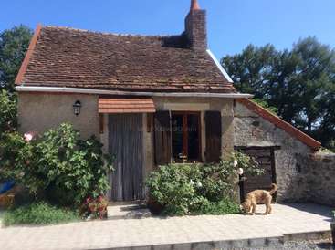 L'image représente une charmante maison en pierre au toit de tuiles brunes, avec une cheminée, une porte et une fenêtre en bois, entourée d'une végétation luxuriante et d'un chien sous le porche.