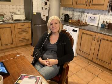 L'image représente une femme assise dans une cuisine, entourée d'armoires et de comptoirs en bois, avec une tablette et un cordon blanc sur la table devant elle.