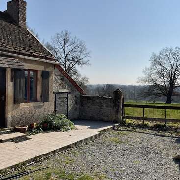L'image représente une charmante maison en pierre avec une cheminée, située sur une allée de gravier, entourée d'une clôture rustique et située dans un cadre rural pittoresque.