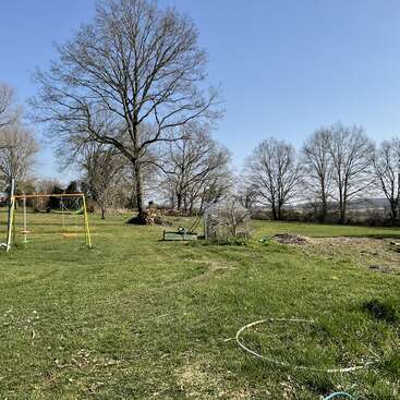 L'image représente un champ herbeux serein avec une balançoire, un banc et un arbre en arrière-plan, sous un ciel bleu clair.