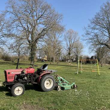 L'image représente un tracteur rouge garé dans un champ herbeux, accompagné d'une tondeuse à gazon verte et d'une balançoire jaune à l'arrière-plan, entourée d'arbres.