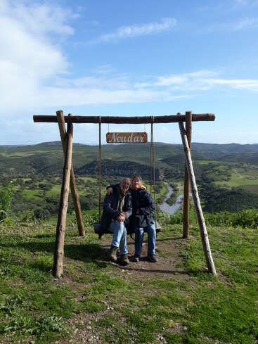 Una pareja se sienta junta en un columpio de madera con un cartel de "Noudar", con vistas a un paisaje pintoresco con colinas verdes, río y un cielo azul brillante.