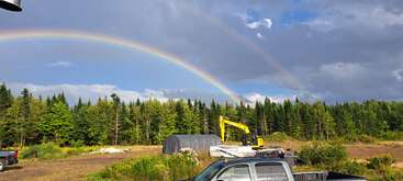 Um arco-íris duplo vibrante se estende por um céu nublado sobre uma floresta. Abaixo, veículos de construção, vegetação e uma estrutura coberta por lona acrescentam interesse à cena rural.