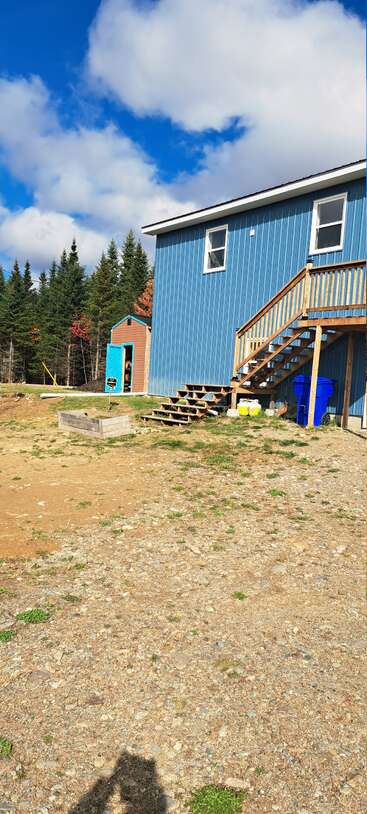 Uma casa azul com escada de madeira, pequeno galpão, lixeiras de reciclagem, jardineira e pátio de cascalho. Cercada por pinheiros, sob um céu azul com nuvens.