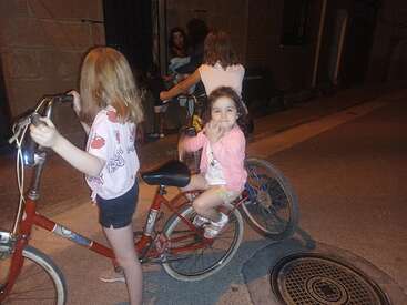 Três meninas estão andando de bicicleta à noite em uma rua tranquila. Uma delas sorri e acena para a câmera, criando uma atmosfera alegre e amigável.