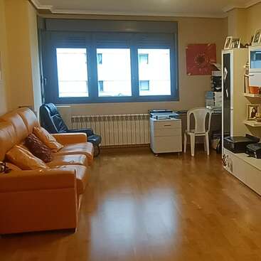 Una sala de estar ordenada con suelo de madera, sofá naranja, silla negra, ventanas grandes, escritorio con ordenador, silla de plástico, estanterías, impresora y un cuadro con flores.