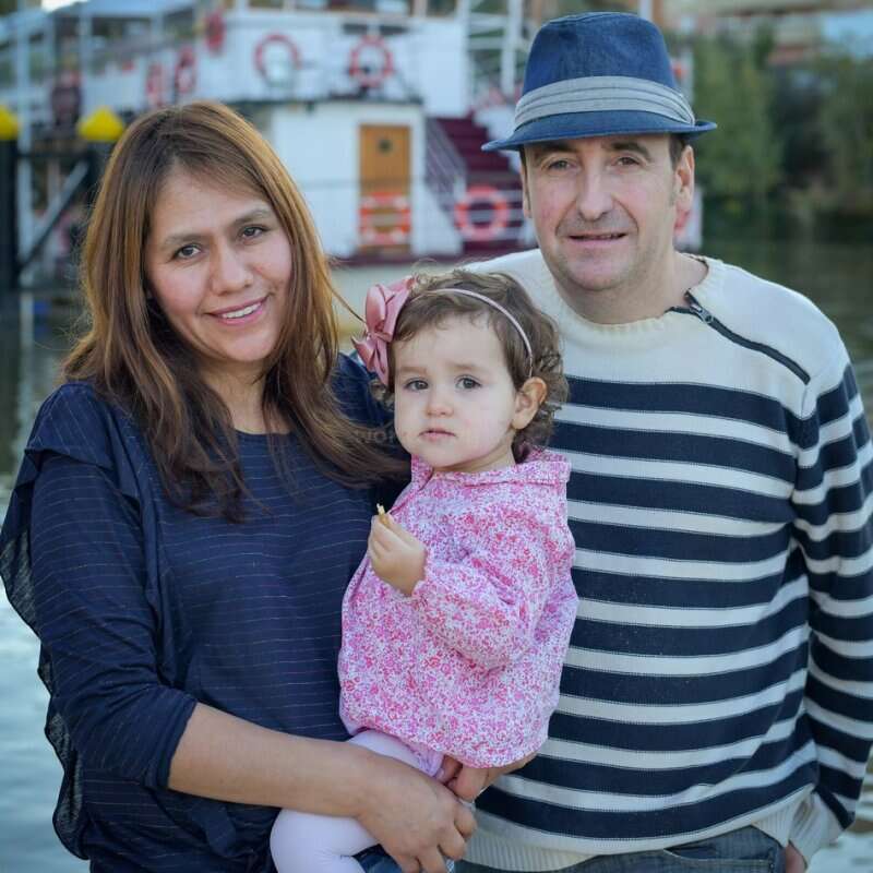 Um casal sorridente posa com sua filha pequena, que usa uma jaqueta rosa com estampa floral e um laço. Eles estão ao ar livre, perto de um barco, curtindo um tempo em família.