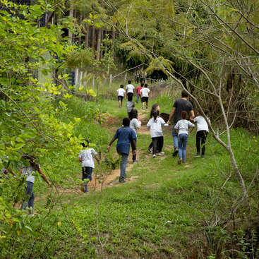 The image depicts a group of children and an adult walking along a dirt path in a wooded area, surrounded by lush greenery and trees.